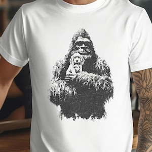 May include: White t-shirt with a black and white illustration of a Bigfoot figure cradling a puppy. The graphic is detailed, showing the Bigfoot's fur and the puppy's features. The t-shirt has a crew neck and short sleeves.