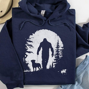 May include: A navy blue hooded sweatshirt featuring a white graphic print of a silhouette of Bigfoot walking a dog on a leash. The design is set against a circular background depicting a forest of pine trees. The hoodie has a drawstring hood.