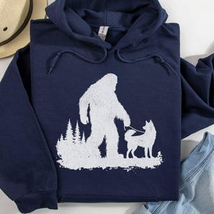 May include: A navy blue hooded sweatshirt featuring a white graphic print of a silhouette of Bigfoot walking a dog on a lead in front of a forest of pine trees. The hoodie has a drawstring hood and long sleeves, with a Gildan brand tag visible.