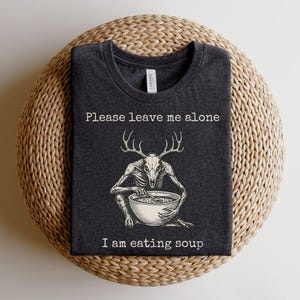 May include: Dark grey t-shirt with the text "Please leave me alone, I am eating soup." The graphic features a skeletal figure with deer antlers, seated and stirring soup in a bowl. The shirt is folded on a woven, circular surface.