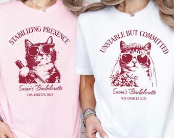 May include: Two t-shirts, one soft pink and one white, featuring a cat wearing sunglasses. The pink shirt reads "STABILIZING PRESENCE" and the white shirt reads "UNSTABLE BUT COMMITTED". Both shirts have "Susan's Bachelorette, Los Angeles 2025" printed below the cat graphic.