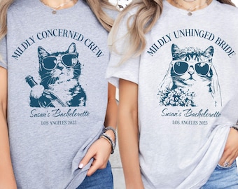 May include: Two light gray t-shirts with matching cat graphics wearing sunglasses. One shirt reads "MILDLY CONCERNED CREW" and the other reads "MILDLY UNHINGED BRIDE". Both shirts have "Susan's Bachelorette LOS ANGELES 2025" printed below the cat.