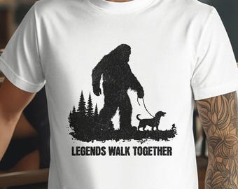 May include: White t-shirt with a black graphic depicting Bigfoot walking a dog. The design includes a forest silhouette and the text "LEGENDS WALK TOGETHER". A casual tee for everyday wear.