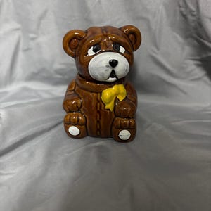 May include: A brown ceramic teddy bear figurine with a yellow bow tie. The bear has white painted eyes and a white nose. The bear is sitting upright with its paws together.