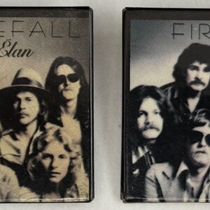 May include: Two vintage cassette tapes featuring the album cover of "Firefall Elan." The black and white image shows a group of men with long hair, sunglasses, and hats. The band's name and album title are printed at the top.