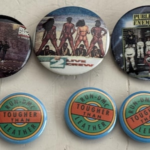 May include: A collection of six round pinback buttons featuring music-related designs. The buttons display images of Run-DMC, 2 Live Crew, and Public Enemy. The Run-DMC buttons have a blue border with the text "Tougher Than Leather".