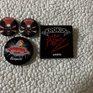May include: Four black and red Krokus band pins. Two pins feature a skull and crossbones design. One pin features the band's name and the text "Europe 81". The fourth pin is a black and red album cover with the text "Krokus The Blitz" and "Arista".