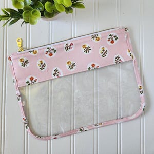 May include: A clear, rectangular cosmetic bag with a pink floral pattern and a gold zipper. The bag features a clear front panel and a pink fabric border with a repeating pattern of white ovals containing yellow and red roses.