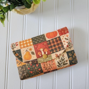 May include: A rectangular fabric pouch featuring a patchwork design of autumn-themed squares. The pattern includes illustrations of orange pumpkins, red mushrooms, pears, and foliage in shades of brown, green, and cream, secured with a gold snap.