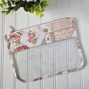 May include: A rectangular zippered pouch featuring a clear plastic lower section and a fabric upper section. The fabric displays a patchwork pattern of pink roses, green leaves, and small floral prints on a white background with a gold metal zipper.