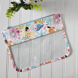 May include: A rectangular clear vinyl pouch featuring a colorful summer-themed print on the upper section. The pattern includes illustrations of a person on a towel, a sun hat, a flamingo, and tropical foliage in shades of pink, blue, and yellow.