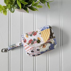 May include: A small fabric pouch featuring a patchwork design with floral patterns in red, white, and blue, alongside yellow striped sections. The pouch has a rounded flap secured with a green button and includes a short strap with a metal swivel clasp.
