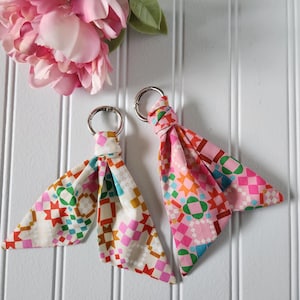 May include: Two fabric keychains featuring a geometric quilt-inspired pattern with stars and squares in shades of pink, green, orange, and blue. Each keychain is attached to a silver-toned metal ring, showcasing a folded, triangular fabric design.