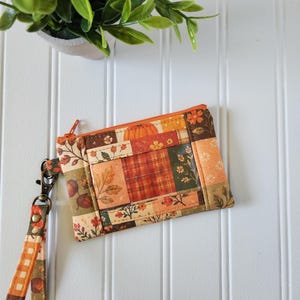 May include: A rectangular fabric wristlet pouch featuring a patchwork design with autumn-themed patterns including pumpkins, acorns, leaves, and plaid in shades of orange, brown, and green. It has an orange zipper and a matching fabric wrist strap.