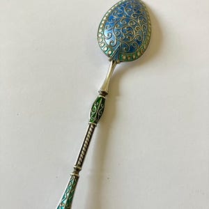 Pode incluir: Uma colher de prata com detalhes em esmalte azul e verde. A colher tem um cabo decorativo com um design torcido e uma tigela arredondada.
