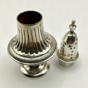 Może przedstawiać: Srebrna solniczka i osobna pieprzniczka. Solniczka ma szeroką, żłobkowaną podstawę i dekoracyjny, żebrowany wzór. Pieprzniczka ma kształt stożka z małymi otworami i zaokrąglonym wierzchołkiem. Oba wykonane są z błyszczącego metalu.