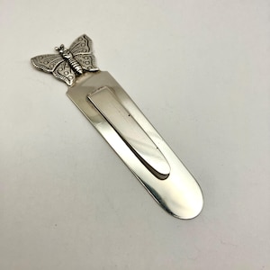 May include: A silver-colored metal bookmark with a butterfly design. The butterfly is at the top of the bookmark, with detailed wing patterns. The bookmark has a curved, clip-like design.