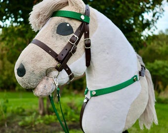 May include: A plush hobby horse toy with a tan and white coat, featuring a light-colored mane and tail. The horse is equipped with a brown leather bridle, silver-toned metal buckles, and bright green nylon straps, all mounted on a wooden stick.