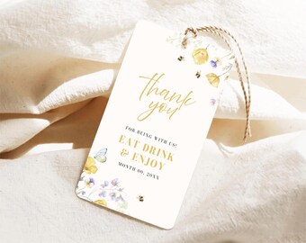 Può includere: Un'etichetta regalo color crema con la scritta dorata "thank you". L'etichetta presenta illustrazioni ad acquerello di fiori gialli e viola, api e una farfalla. L'etichetta recita "EAT DRINK & ENJOY". È attaccato uno spago.