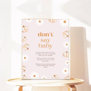 Può includere: Un cartello rettangolare con la scritta "don't say baby" in arancione. Il cartello è decorato con zucche e margherite su sfondo rosa chiaro. È per un gioco di baby shower.