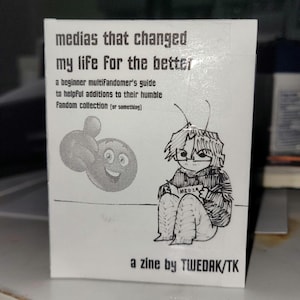 May include: A white zine titled "medias that changed my life for the better." The cover features a cartoon illustration of a person writing and a thumbs-up emoji. Text includes "a beginner multifandomer's guide" and "a zine by TWEDAK/TK."