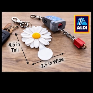 Op de afbeelding: Een witte madeliefjes sleutelhanger met een gele kern, 11,4 cm hoog en 6,4 cm breed, naast een autosleutel en een afstandsbediening. De sleutelhanger is bevestigd aan een zilveren sluiting. Het Aldi-logo is zichtbaar.