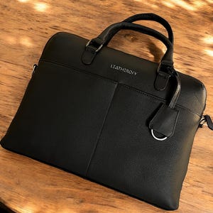 May include: A black briefcase with a textured finish, featuring two top handles and a silver-toned metal ring attachment. The word LEATHEROFY is printed in silver lettering on the front. The bag has a structured rectangular shape with clean lines.