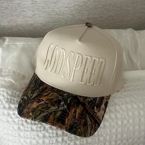 May include: A beige baseball cap with the embroidered text "GODSPEED" in a matching color. The brim features a camouflage pattern in shades of brown, green, and black. The cap is resting on a white pillow.