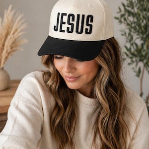 May include: A cream-colored baseball cap with a black brim and a black button on top. The word "JESUS" is embroidered in bold, black letters across the front of the cap. The cap is worn by a person.