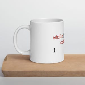 Könnte beinhalten: Weiße Keramiktasse mit schwarzem Text, der "while(coding) { }" in roter Schrift lautet.