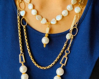 May include: A layered gold chain necklace with a large pearl pendant and a smaller pearl charm. The necklace features a variety of chain styles, including a box chain, a cable chain, and a curb chain. The pearls are white and have a smooth, polished finish.