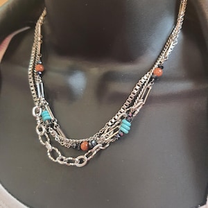 May include: A layered silver chain necklace with turquoise and brown beads. The necklace has a delicate, bohemian style.