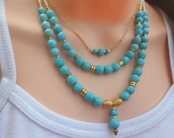 May include: A layered turquoise bead necklace with gold accents. The necklace features three strands of turquoise beads, with a smaller strand of gold beads in the center. The necklace has a gold pendant with a turquoise bead at the bottom.