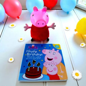 May include: A Peppa Pig soft toy in a red dress is positioned near a "Happy Birthday" book with a cake illustration. Balloons in red, blue, and yellow are in the background, along with daisy cutouts. The book cover features Peppa Pig.