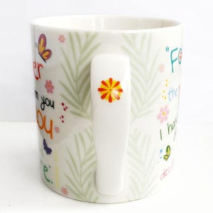 May include: A white ceramic mug featuring a light green fern leaf pattern, colorful butterflies, and small flowers. The handle is decorated with a small orange and yellow flower. The mug displays partial text in various colors and fonts.