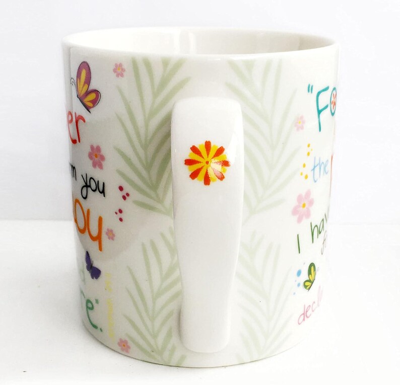 May include: A white ceramic mug featuring a light green fern leaf pattern, colorful butterflies, and small flowers. The handle is decorated with a small orange and yellow flower. The mug displays partial text in various colors and fonts.