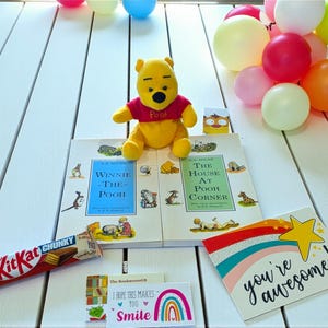 May include: A yellow Winnie the Pooh plush toy sits atop two books, "Winnie-the-Pooh" and "The House at Pooh Corner." A KitKat Chunky bar, a card with a rainbow and the text "you're awesome!", and balloons are also present.