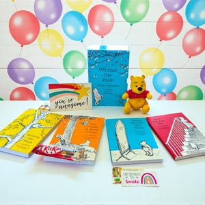 May include: A collection of Winnie-the-Pooh books, a plush toy, and greeting cards are arranged on a white surface. The books have colorful covers, and the plush toy is of Winnie-the-Pooh. A Kit Kat candy bar is also included.