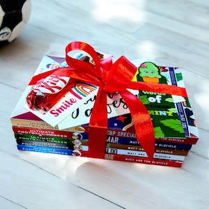 Könnte beinhalten: Ein Stapel von fünf Büchern, die mit einem roten Band umwickelt sind, mit einer Schokoladentafel und einer Karte mit der Aufschrift "Lächeln, du bist großartig". Die Bücher handeln vom Fußball und haben Titel wie "Ultimate Football Heroes" und "Cup Special - Matt and Tom Glofield".