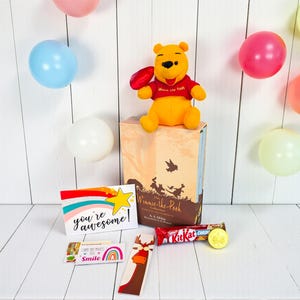 May include: A Winnie the Pooh themed gift set with a plush toy, book, greeting cards, Kit Kat bar, and a gold coin. Colorful balloons in pink, blue, and white are in the background. The greeting card says "you're awesome!"