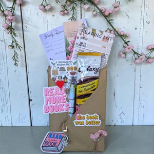 May include: A brown paper envelope containing a book review sheet, a reading list, a Galaxy caramel hot chocolate packet, a patterned pen, and stickers reading Read More Books, The Book Was Better, and Read A Damn Book with a heart clip.