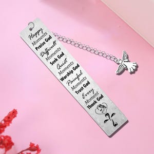 May include: A rectangular silver-toned metal bookmark featuring a chain with an angel charm. The surface is engraved with the text: Happy Moments Praise God, Difficult Moments Seek God, Quiet Moments Worship God, Painful Moments Trust God, Every Moment Thank God.