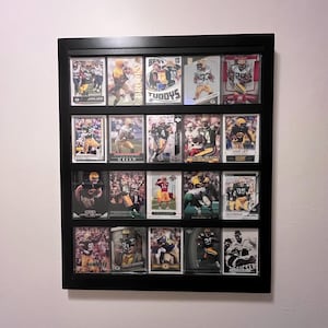 May include: A black framed display case filled with football trading cards. The cards feature players in green and yellow uniforms. The case is mounted on a white wall.