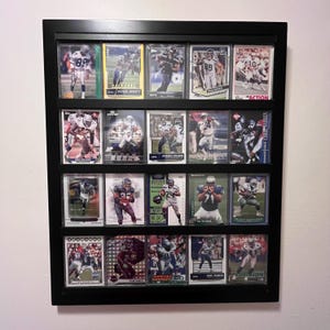 May include: A black rectangular wall-mounted display case containing twenty individual American football trading cards arranged in four horizontal rows. Each card features a player in uniform, with various team colors and action poses visible.