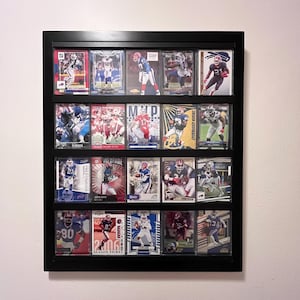May include: A black rectangular wall-mounted display case containing twenty individual sports trading cards arranged in four rows of five. Each card features a football player in uniform, with various team colors, action poses, and printed text visible.