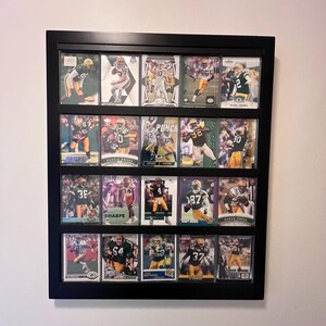 May include: A black rectangular wall-mounted display case containing twenty individual trading cards arranged in four rows of five. Each card features a football player in a green and yellow uniform, protected by clear plastic sleeves within the frame.