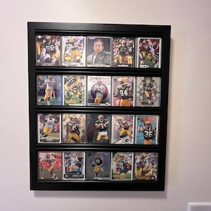 May include: A black framed display case filled with football trading cards. The cards feature players in green and yellow uniforms. The case is mounted on a white wall, showcasing a collection of sports memorabilia.
