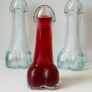 May include: Three clear glass bottles shaped like a phallus. One bottle is filled with a red liquid. The bottles are on a white surface. The bottles have a bulbous base and a rounded top. The image includes the text "@NaughtThatDeep".