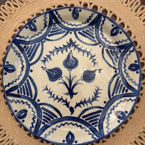 May include: A round ceramic plate with a cream-colored base and a speckled finish. The plate features a hand-painted dark blue pattern consisting of a central floral motif surrounded by repeating arched geometric shapes and decorative lines.