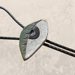 May include: A ceramic bolo tie featuring a stylized eye design with green, white, and black accents. The bolo tie is attached to a black cord.
